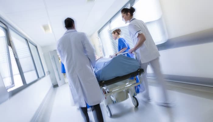 A motion blurred photograph of a patient on stretcher or gurney being pushed at speed through a hospital corridor by doctors & nurses to an emergency room.
